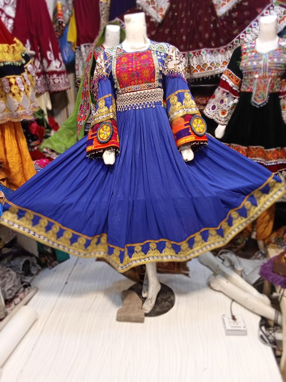 “Mannequin dressed in a vibrant blue traditional Afghani frock featuring intricate embroidery, surrounded by ethnic garments that highlight Afghani embroidery and gemstone jewelry craftsmanship.”