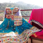 A traditional Afghan dress in deep blue with intricate multicolored embroidery, paired with a vibrant pink shawl featuring decorative borders. The outfit is displayed on a chair placed on a balcony, with a scenic view of green trees and distant mountains in the background. This image reflects Afghan embroidery storytelling, where each stitch conveys cultural heritage and artisanal elegance.