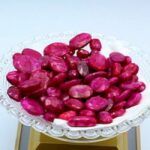 "A cluster of polished ruby stones in varying shapes and sizes, displayed on a scalloped white ceramic plate with a vibrant red gloss. The plate rests on a yellow stand, highlighting the deep crimson hues and lustrous finish of the gemstones."