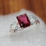 A close-up of a ruby ring featuring a vibrant, square-cut ruby set in rhodium-plated silver. The band showcases elegant detailing, and the ring rests on a soft white cushion with a warm wooden background.