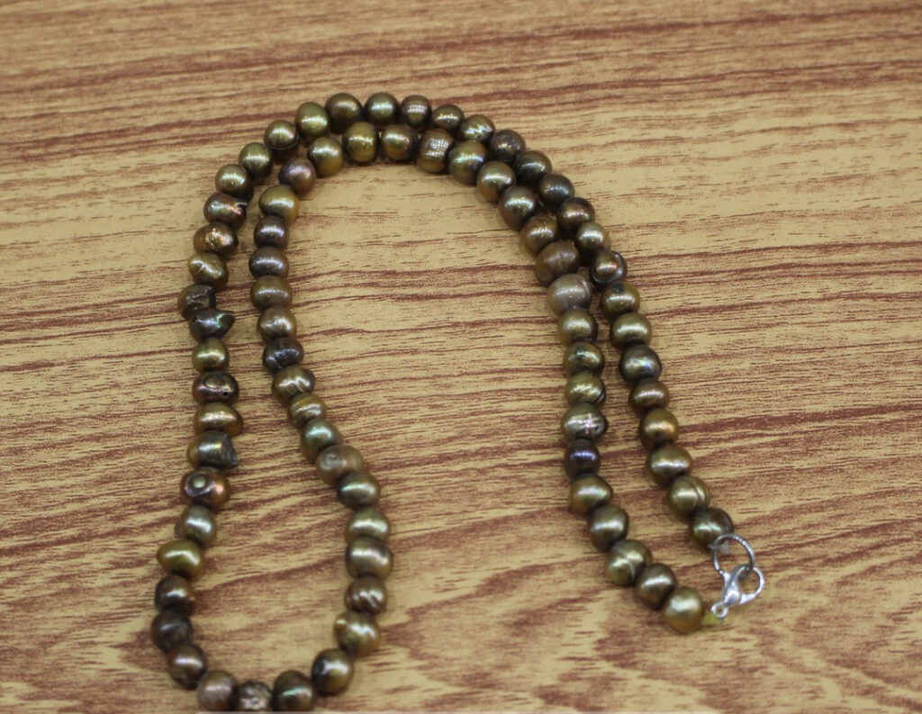 A handcrafted necklace of dark, naturally irregular lustrous pearls, elegantly arranged on a warm-toned wooden surface. Each pearl glows with a metallic sheen and organic charm, reflecting artisanal beauty and earthy sophistication.