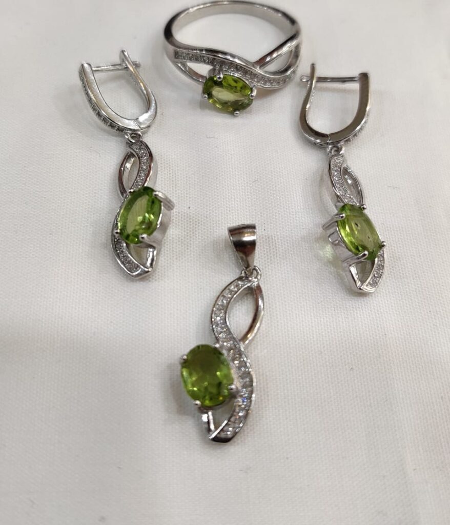 Silver peridot jewelry set displayed on soft white fabric, including a ring, pendant, and earrings with green gemstones.