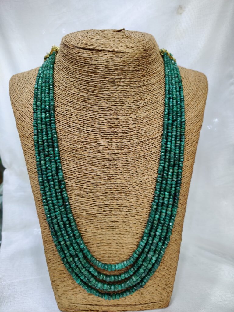 Multi-strand emerald necklace displayed on a woven, textured stand. The necklace features several rows of small, polished emerald beads in rich green tones, arranged in cascading layers that evoke elegance and vitality.