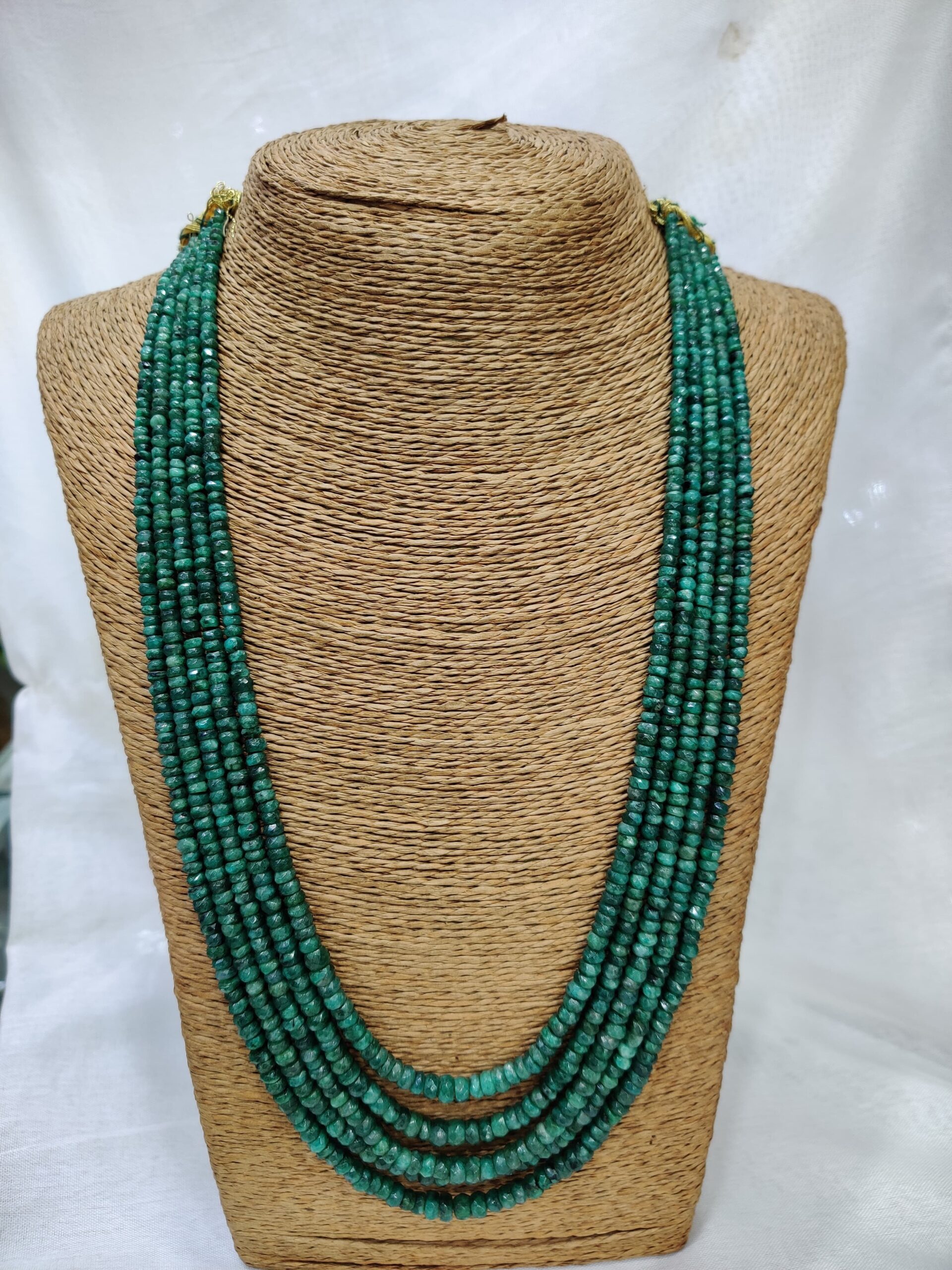 Multi-strand emerald necklace displayed on a woven, textured stand. The necklace features several rows of small, polished emerald beads in rich green tones, arranged in cascading layers that evoke elegance and vitality.