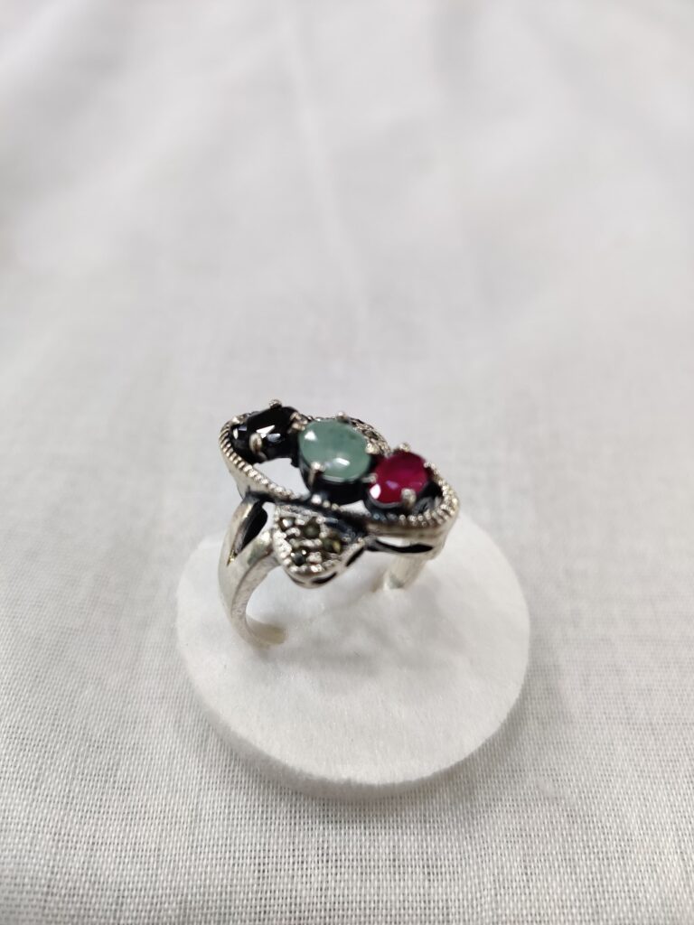 Silver ring with green and red gemstones, elegantly set in an intricate design, displayed atop a white circular stand against a soft gray background. The account page