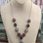 "Silver necklace from our gemstones collection with red, green, and blue gemstone clusters arranged in a V-shape, displayed on a beige mannequin bust in a jewelry store."