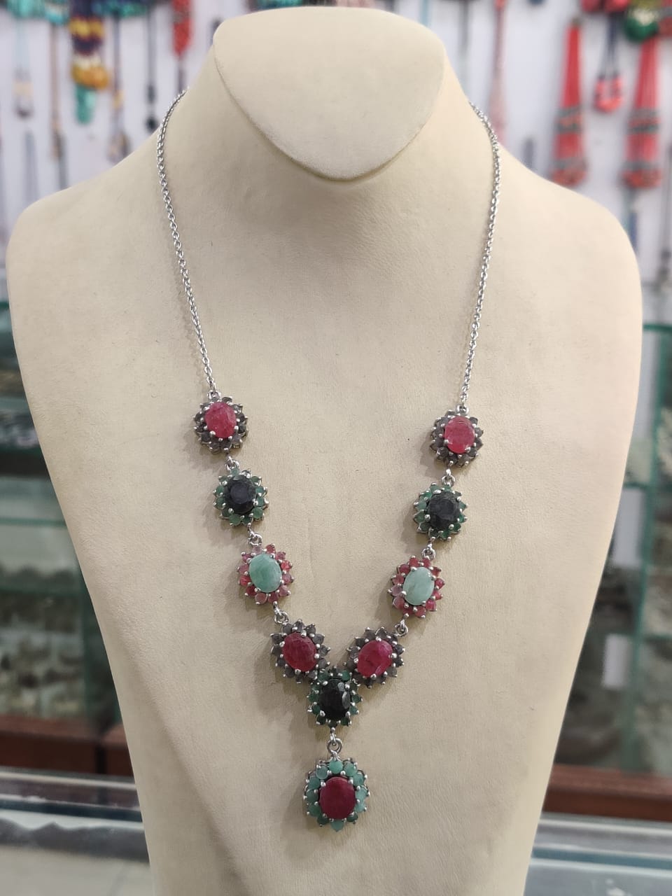 "Silver necklace from our gemstones collection with red, green, and blue gemstone clusters arranged in a V-shape, displayed on a beige mannequin bust in a jewelry store."